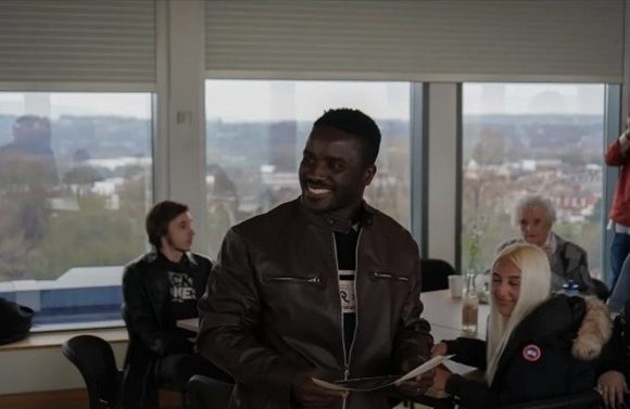 Person standing and holding papers in a meeting room with large windows overlooking a town or city. Several other people are seated around tables in the background, along with drinks and bags, in a bright indoor setting.